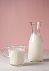 A glass of milk is next to a milk jug. pink background