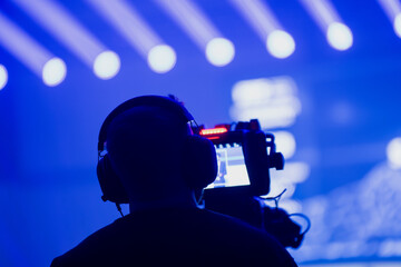 Camera operator filming a live event under intense blue stage lighting with bright beam patterns, showcasing professional broadcast and event production workflow
