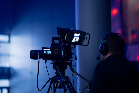 Broadcast camera operator filming a live indoor event using a professional tripod-mounted video camera with monitoring screens and headset communication