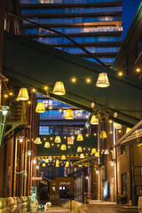 Fototapeta premium Vertical view of a decorated alley in Torontos Distillery District at night with hanging lamps and warm string lights between historic brick buildings.
