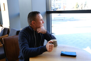 an adult man sits by the window leaning on his arm man drinking a coffee in a coffee shop with a laptop and headphones on the table. High quality photo