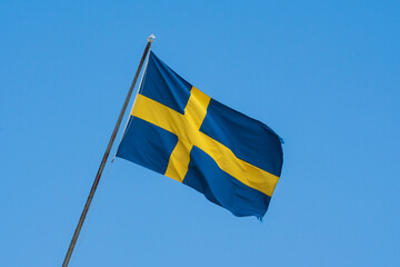 Waving national flag of Sweden with yellow cross on blue background flying on a flagpole against clear sky