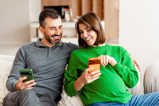 Pregnant couple relaxing on couch using smartphones sitting on comfortable sofa at home