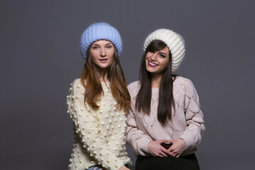 Portrait of Female Friends with Contrasting Expressions, One Smiling Broadly and One with a Soft Expression, Both Wearing Cozy Woolen Hats and Sweaters in a Studio Setting
