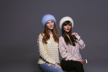Waist-Up Shot of Two Female Models Displaying Warm Autumn or Winter Knitwear, Featuring a Blue Mohair Hat and Off-Shoulder Yellow Sweater Next to a White Ribbed Beanie and Patterned Beige Pullover