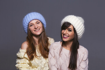 Portrait of Female Friends with Contrasting Expressions, One Smiling Broadly and One with a Soft Expression, Both Wearing Cozy Woolen Hats and Sweaters in a Studio Setting