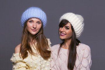 Portrait of Female Friends with Contrasting Expressions, One Smiling Broadly and One with a Soft Expression, Both Wearing Cozy Woolen Hats and Sweaters in a Studio Setting