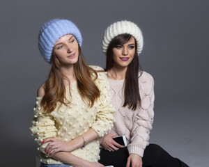 Waist-Up Shot of Two Female Models Displaying Warm Autumn or Winter Knitwear, Featuring a Blue Mohair Hat and Off-Shoulder Yellow Sweater Next to a White Ribbed Beanie and Patterned Beige Pullover