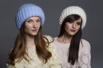 Waist-Up Shot of Two Female Models Displaying Warm Autumn or Winter Knitwear, Featuring a Blue Mohair Hat and Off-Shoulder Yellow Sweater Next to a White Ribbed Beanie and Patterned Beige Pullover