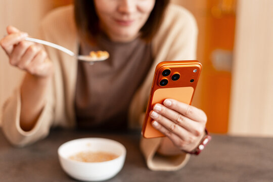 Woman eating breakfast cereal while browsing smartphone selective focus on hands