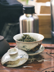 A ceramic gaiwan containing dry green tea leaves sits open on a carved wooden tray, ready for brewing in a traditional Chinese gongfu tea session.