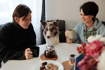 Two women sit at a bright kitchen table with a border collie between them, enjoying warm drinks in a peaceful morning atmosphere. Companionship, shared routine, and gentle daily harmony with a pet.