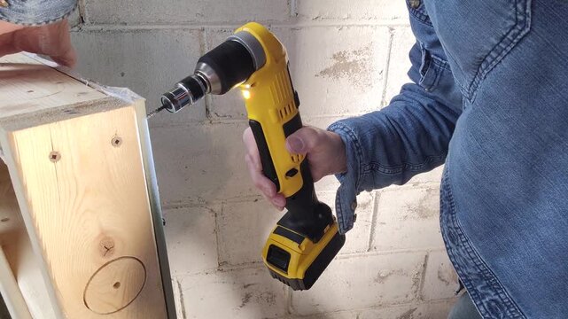 A worker uses a battery operated cordless right angle drill to drive screws into drywall with a magnetic screw driver attachment