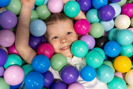 Happy smiling boy lying in colorful ball pit with raised arms at indoor playground looking at camera. Concept of children entertainment, kids amusement center fun and joyful childhood moments. - Powered by Adobe
