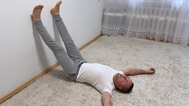 A grown man lies on the floor on a carpet at home with his legs raised up on the wall and his arms spread out to the sides. High quality photo