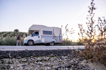 A bearded man stands in front of his camper van, gesturing in frustration while speaking on the phone. The open hood and rural surroundings emphasize the unexpected breakdown.