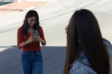 Smiling young woman happily photographing a friend with her smartphone, capturing moments of friendship and sharing cheerful experiences outdoors on the street