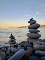 Two carefully balanced stacks of smooth, grey stones (cairns) stand on a pebble beach by the calm sea, symbolizing balance, meditation, or tranquility.