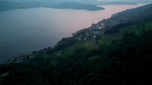 Cinematic aerial footage during blue hour showing a Swiss lakeside town with warm lights, calm water and mountains in the background. Soft pink sky and a tranquil atmosphere. Ideal for storytelling, t