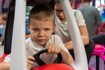 Focused little boy driving red toy car with serious expression while older brother watches at indoor playground. Concept of children entertainment, siblings play, pretend driving and amusement center.