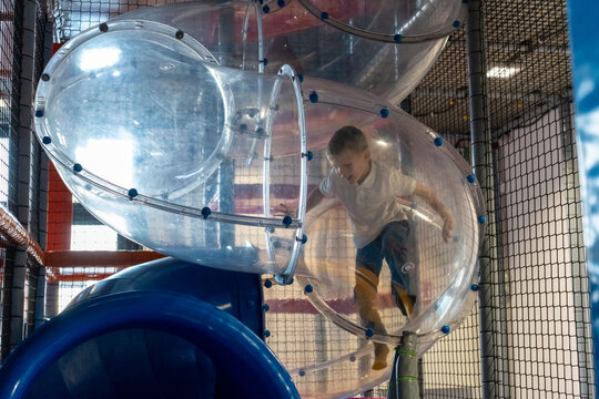 Boy climbing inside transparent bubble tunnel connecting play structures at indoor playground with blue slide and safety nets. Concept of children adventure, exploration play and amusement center fun.