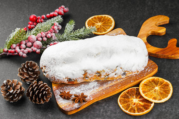 Classic Christmas Stollen covered with powdered sugar icing on wooden board with dried oranges, pine branches and cones on black textured surface. Traditional holiday food, sweet fruit loaf