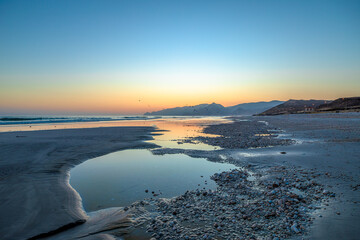 Wild seagulls gliding and settling on the wet sand of Mughsayl Beach near Salalah in Dhofar, Oman, as waves roll ashore and distant mountains appear in silhouette under a colorful dry-season sunset