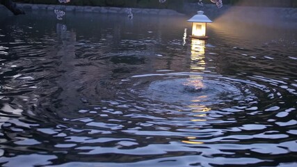 Cherry blossoms hanging over a traditional japanese garden pond at twilight, a stone lantern illuminating the dark water and creating ripples from a small fountain - Powered by Adobe