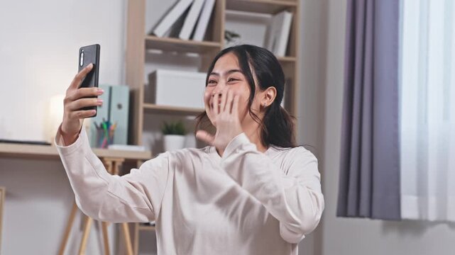 Asian Young woman holding smartphone taking frustrated video call selfie cozy home office with shelves and soft lighting while later smiling naturally and showing relaxed confident expression during - Powered by Adobe