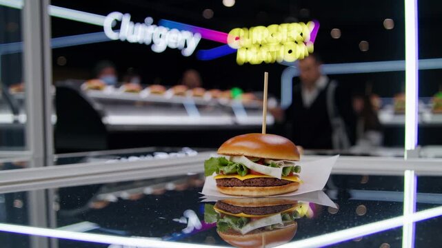 cheeseburgers are neatly arranged on a reflective surface, possibly in a contemporary burger restaurant, with blurred figures of restaurant staff in the background