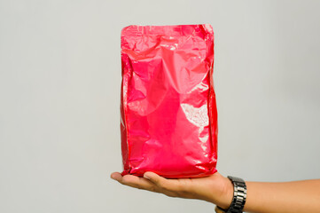 A person's hand in a black shirt holds up an aluminum foil pouch, a blank red product packaging bag, which can be used for coffee, snacks, or powder, allowing for easy product mockup and branding.