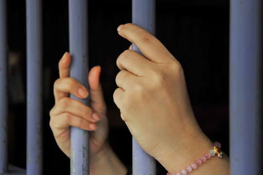 A pair of hands reaching through vertical steel bars of a jail or prison cell, with one hand wearing a pink beaded bracelet, symbolizing confinement, hope, despair, and freedom lost.