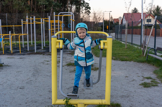 Smiling little boy in blue helmet and colorful jacket using outdoor elliptical trainer at public fitness playground in suburban area at dusk. Concept of early sports education and active childhood.