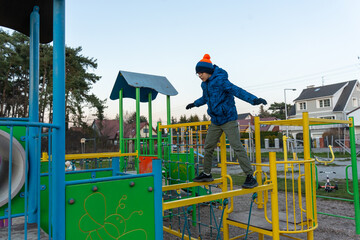 Boy in blue winter jacket balancing and walking on colorful playground climbing structure in suburban neighborhood on cold autumn day. Concept of outdoor play, balance training, active childhood fun.