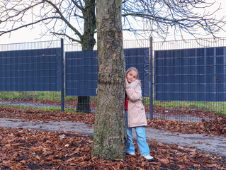Young girl with light brown hair wearing a beige coat is playfully hiding behind a tree in a park surrounded by fallen leaves and a serene atmosphere