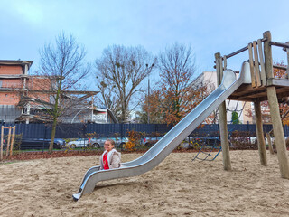 Young girl with blonde hair sitting on a playground slide, surrounded by trees and sand, enjoying outdoor playtime in a vibrant park setting with clear blue sky