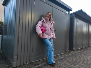 Young woman with long blonde hair wearing a pink puffer jacket and blue jeans, leaning against a gray storage shed while holding a pink soccer ball, showcasing a casual outdoor lifestyle