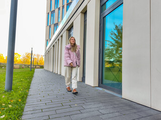 Young woman with long hair wearing a pink jacket walks along a modern building's sidewalk, surrounded by green grass and trees, showcasing urban lifestyle and fashion