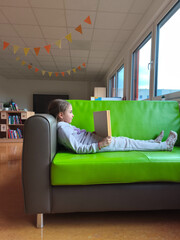 Young girl with brown hair, wearing gray pajamas, is reading a book while lounging on a vibrant green sofa in a cozy room with colorful decorations and natural light