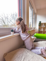 Young girl in cozy pajamas gazes out the window, resting her chin on her hands, surrounded by soft cushions and a small plant, capturing a moment of contemplation and wonder
