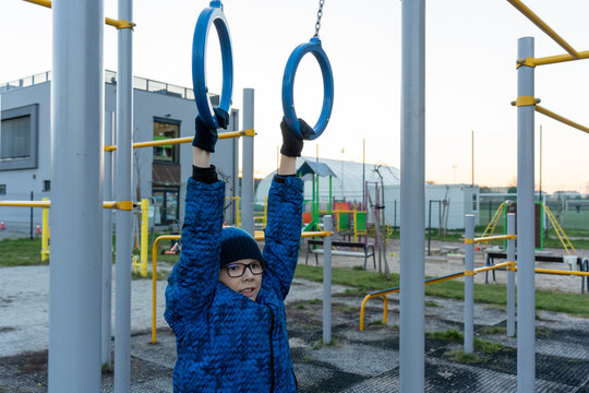 Boy in blue jacket and glasses hanging on gymnastic rings at outdoor fitness playground in suburban area at sunset. Concept of children exercise, street workout, active childhood, physical education. - Powered by Adobe
