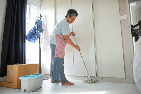 Asian senior woman cleaning floor with mop in bright living room, Elderly lady doing household chores and mopping the floor at home, Active senior woman maintaining a clean home interior