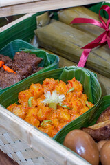 Close up of Indonesian kentang balado, bacem eggs, bacem tempeh, rendang served and lontong in banana leaf containers inside a woven bamboo basket