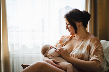 A mother sits in a wooden chair, gently breastfeeding her baby in a warm and softly lit room. Sunlight filters through the curtains, creating a peaceful atmosphere.