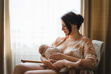 A mother sits in a wooden chair, gently breastfeeding her baby in a warm and softly lit room. Sunlight filters through the curtains, creating a peaceful atmosphere.