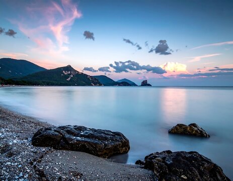 Calm ocean scene at dusk with mountains, rocks, soft clouds, and pastel-colored sky - Powered by Adobe