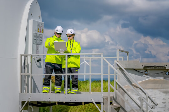 Two workers check data on a laptop at a wind turbine site under a cloudy sky