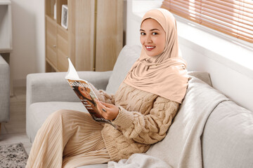 Young Muslim woman reading magazine on sofa at home