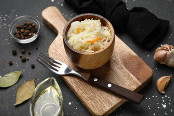 Wooden board with bowl of delicious sauerkraut and different spices on black background