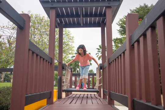 Happy Asian girl balancing and walking across a wooden rope bridge in the outdoor park playground at sunset, enjoying active fun